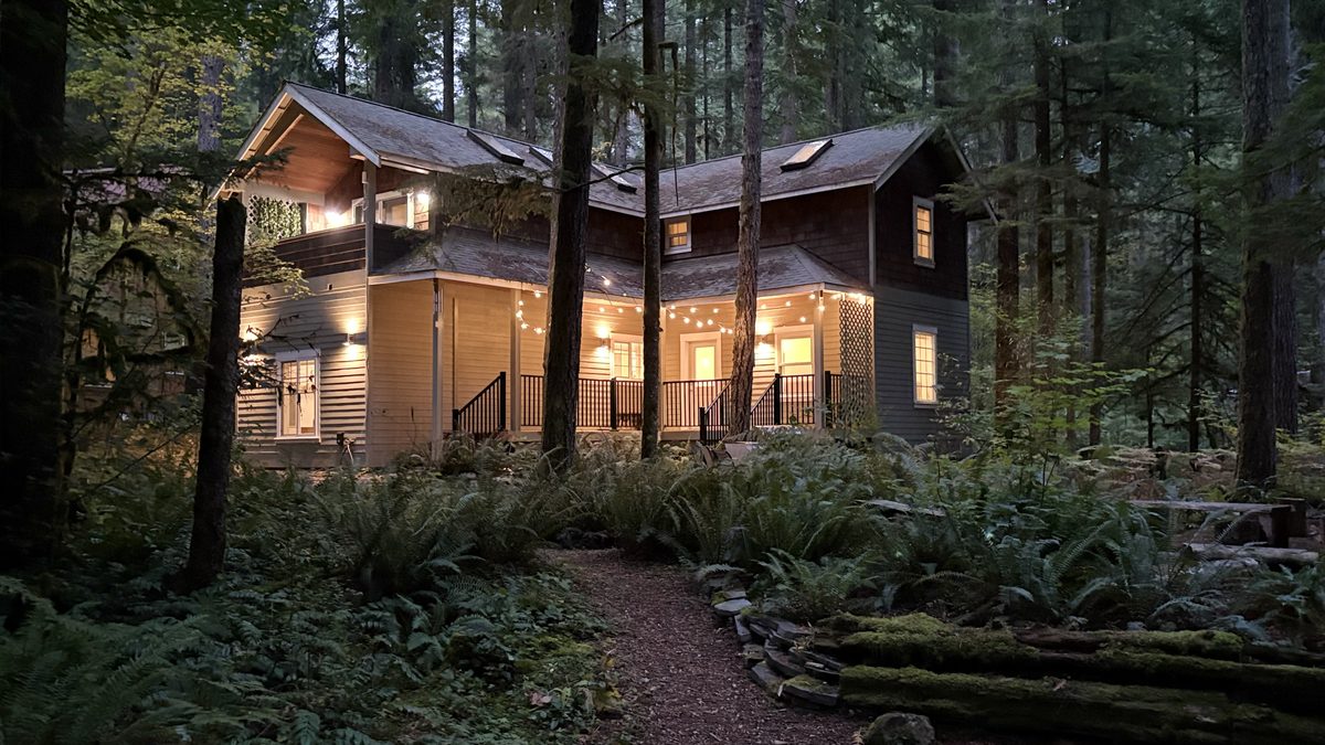 The cabin at dusk with warm windows and string lights among dark firs