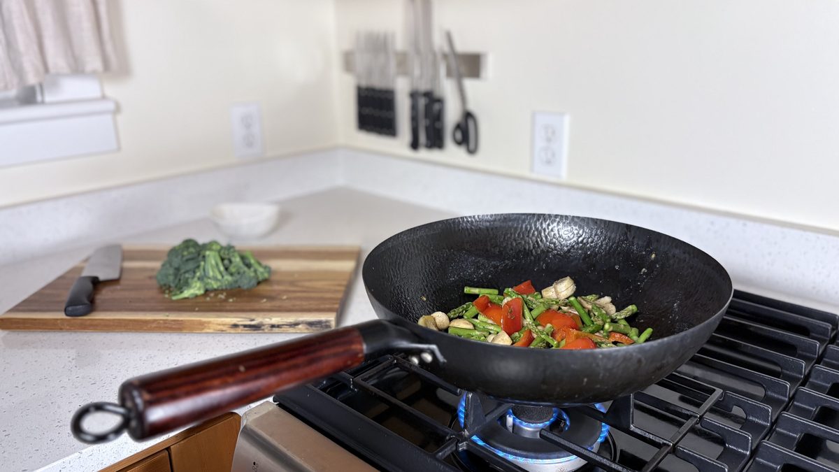 Vegetables stir-frying in a carbon-steel wok on the gas burner, broccoli on a cutting board and knives on a magnetic strip